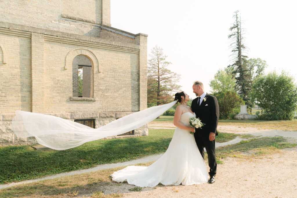 Fine art wedding couple with flowing cathedral veil in front of St Norbert Monastery architecture in Winnipeg with soft natural light