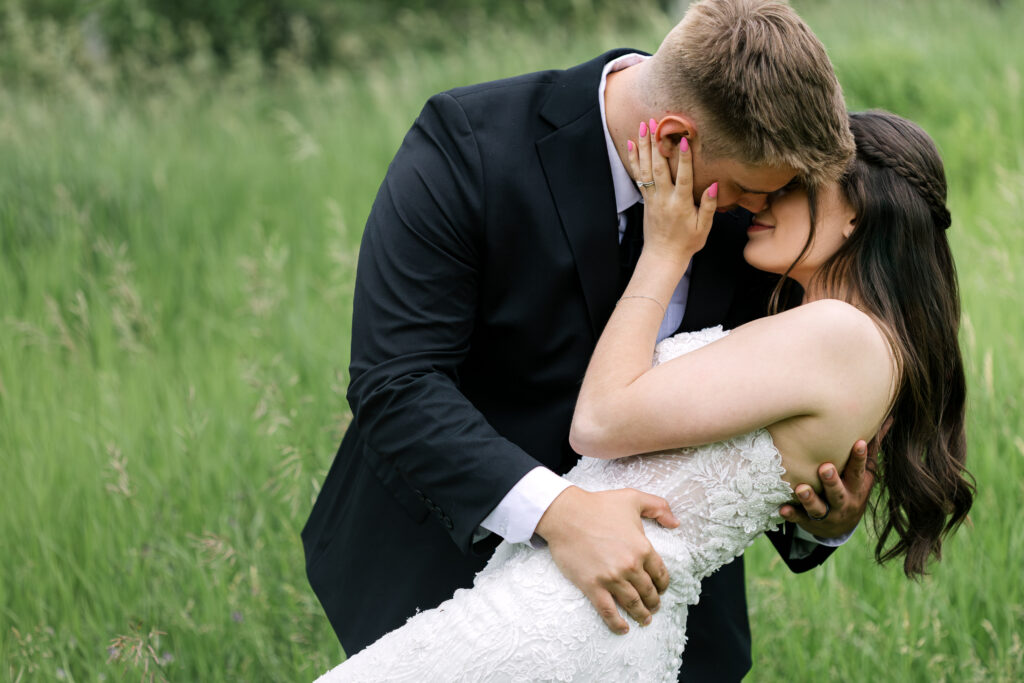Bride and groom sharing a romantic dip and kiss in a grassy field during wedding day in Winnipeg Manitoba with soft natural light
