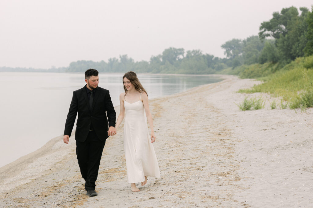 Bride and groom walking along a shoreline during wedding photos in Winnipeg Manitoba with soft natural light and overcast sky